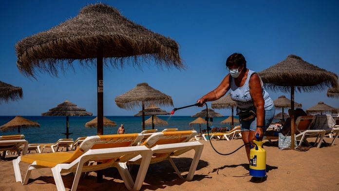 A worker wearing a protective face mask sprays disinfectant on sun loungers at Quarteira Beach in Quarteira, Algarve region, Portugal | Bloomberg