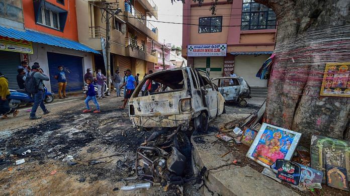 Residents walk past charred remains of vehicles vandalised by a mob over a social media post, allegedly by a relative of a Congress MLA, in Bengaluru, Wednesday, 12 August, 2020.| PTI