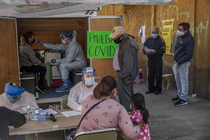 A medical worker wearing protective gear tests a resident while people stand in line at a temporary Covid-19 testing kiosk in the Pedregal de Santo Domingo, Mexico | Photographer: Alejandro Cegarra | Bloomberg