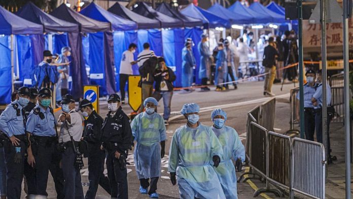 Government workers wearing personal protective equipment (PPE) walk past police officers in an area under lockdown on Robinson Road at night in the Mid-Levels neighborhood of Hong Kong, China, on Saturday, March 13, 2021. | Photographer: Justin Chin | Bloomberg