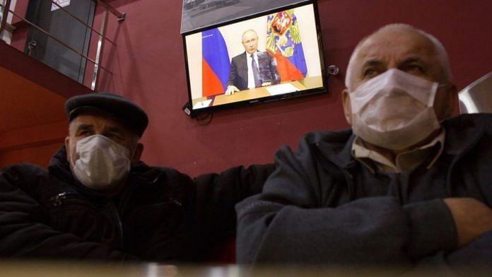 Customers wear protective face masks inside a cafe as a television screen displays Russian President Vladimir Putin delivering a national address, on 25 March 2020 | Bloomberg
