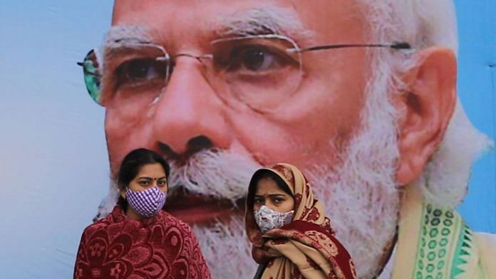 People wearing protective masks stand in front of PM Narendra Modi's posture outside AIIMS in New Delhi | Representational image | Bloomberg