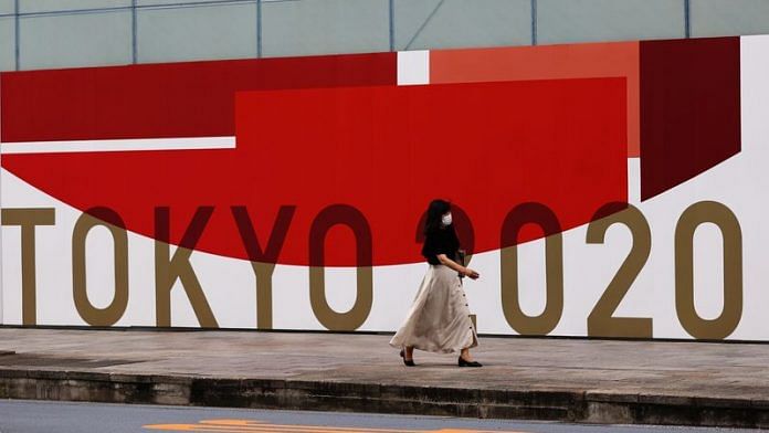 A woman, wearing a protective mask against Covid-19, walks past a wall bearing logo of Tokyo 2020 Olympic Games in Tokyo on 29 July, 2021 | File Photo: Reuters