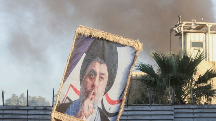 A protester holds up a portrait of Iraqi Shi'ite cleric Moqtada al-Sadr as smoke rises from the Swedish embassy building in Baghdad during a protest near the embassy hours after it was stormed and set on fire ahead of an expected Koran burning in Stockholm, in Baghdad, Iraq, July 20, 2023 | Reuters