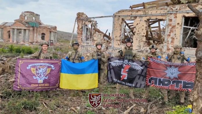 Soldiers hold flags as they speak in front of destroyed buildings in Klishchiivka, Ukraine, as seen in this screen grab taken from a social media video | Handout via Reuters