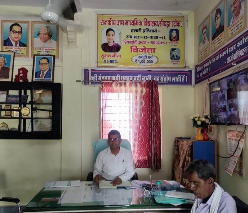 सीदड़ा स्कूल के प्रिंसिपल बीरबल मीणा अपने कार्यालय में. उनके पीछे नेशनल आइकन और स्कूल टॉपर्स के पोस्टर हैं, जिनमें कल्पना मीणा (ऊपर दाईं ओर) शामिल हैं | तस्वीर: कृष्ण मुरारी/दिप्रिंट