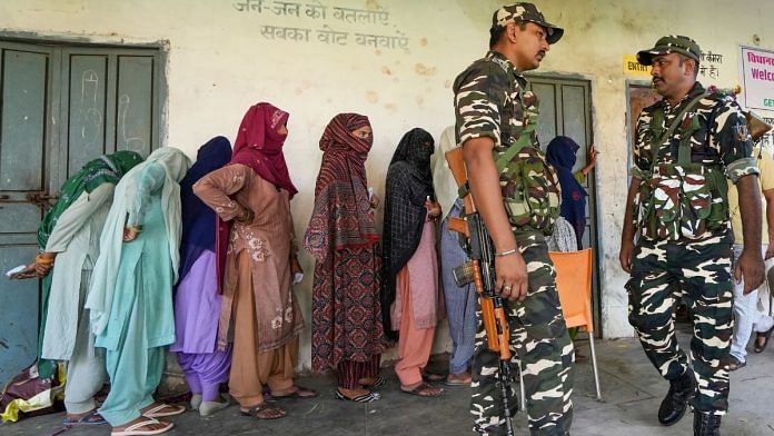 Security personnel guard as people queue up at a polling station to cast their votes for the Haryana Assembly elections, in Nuh district, Saturday, Oct. 5, 2024. (PTI Photo/Kamal Kishore)